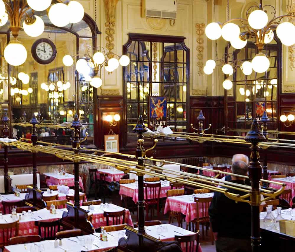 The interior of Restaurant Bouillon Chartier, in Paris, showing its Belle Epoque style, with a high glass ceiling, globe light chandeliers, soaring mirrors and red-and-white tablecloths topped with white paper for writing your order and calculating your bill on.