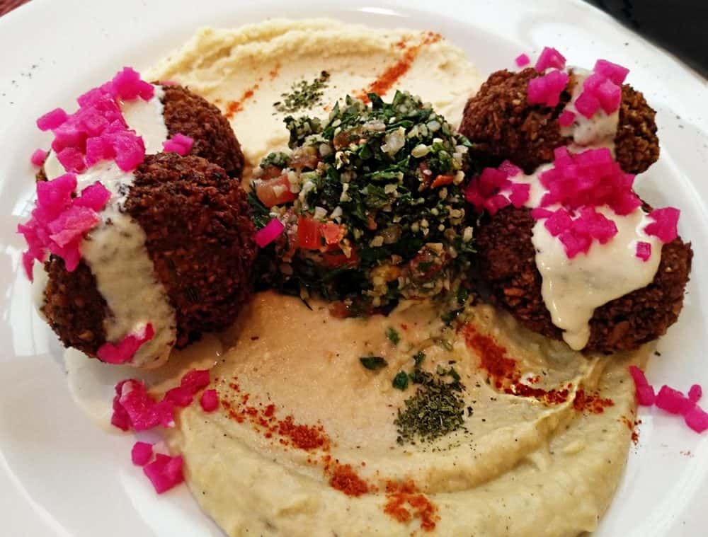 A platter of hummus, falafel, and tabulle topped with cream sauce and bright pink chopped marinated onions.