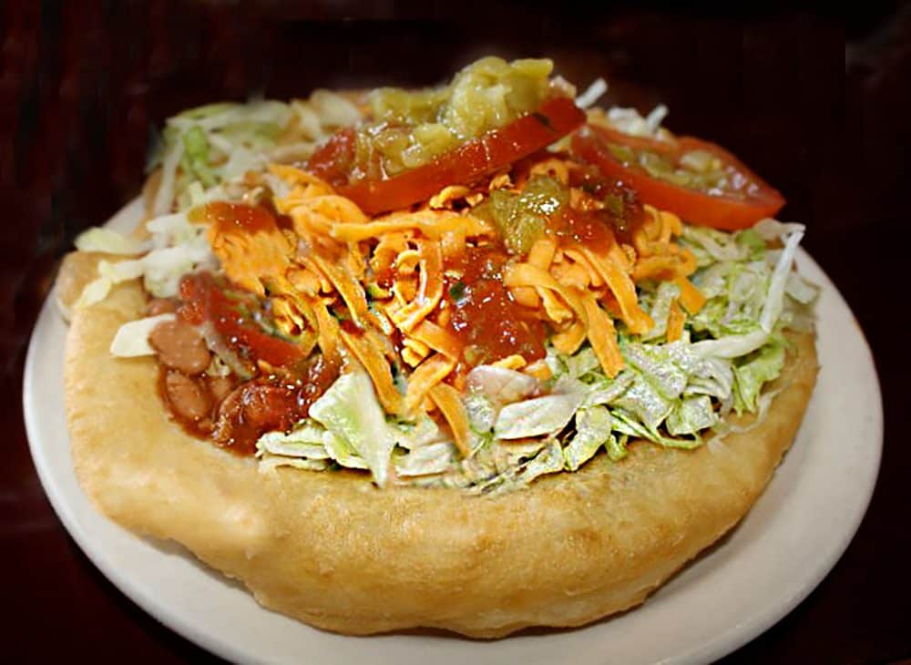 A Navajo Taco, the Cameron Trading Post Restaurant's #1 best seller. This shows a big disk of Indian Fry Bread topped with seasoned ground beef, chile beans, lettuce, tomato, cheddar cheese, and mild green chiles.