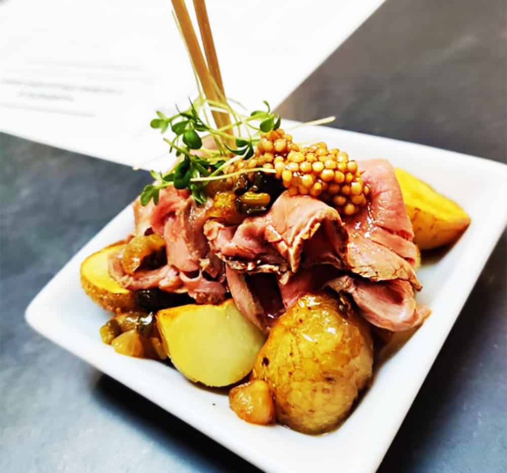 A small plate of sliced pink roast beef folded over mashed potatoes with demi-glace and pickled vegetables, topped with grainy mustard. a two-skewer dish at Bistro Spejle